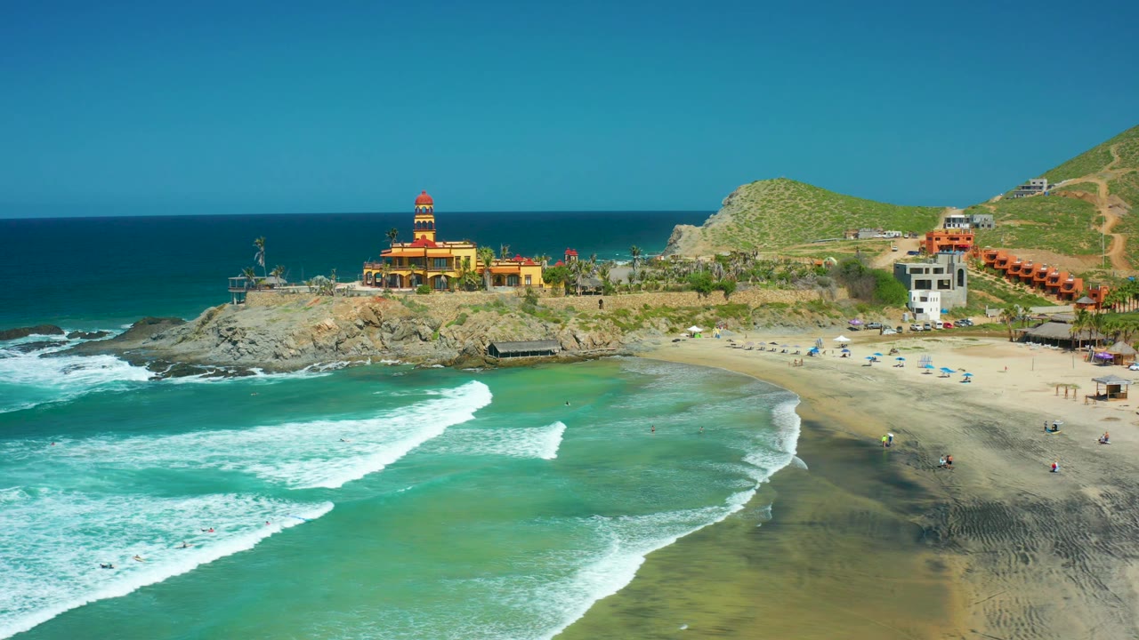 At Cerritos Beach, a broad white wave line advances through green-blue water while people remain small on the beach and the lighthouse-style building sits above the rocks.
