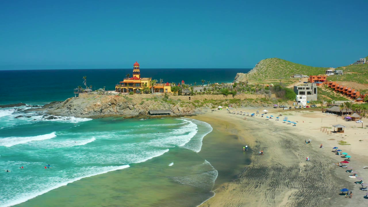 Wide aerial beach view with surf, swimmers, umbrellas, cliffs, and lighthouse.
