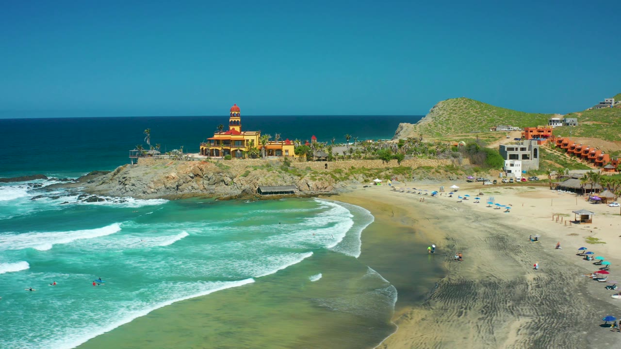 A high oblique view over Cerritos Beach shows a curved white wave line in the foreground, scattered beachgoers near the shore, umbrellas on the sand, and the lighthouse backed by green hills and ocean horizon.