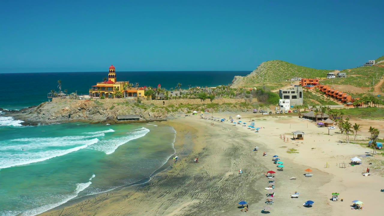Wide aerial beach scene with lighthouse, rocky coast, umbrellas, and rolling surf