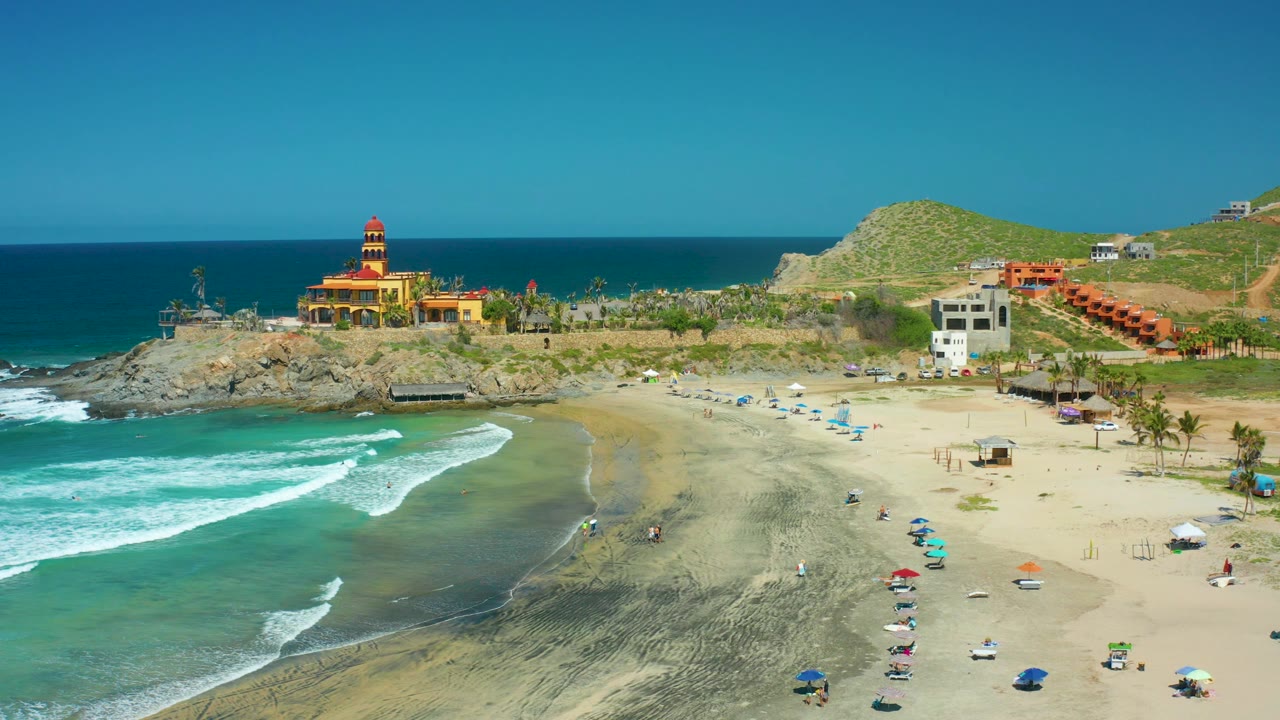 At 0:02, turquoise water fills the lower left while small figures and umbrellas dot the sand beneath the lighthouse and the white and orange buildings on the slope.