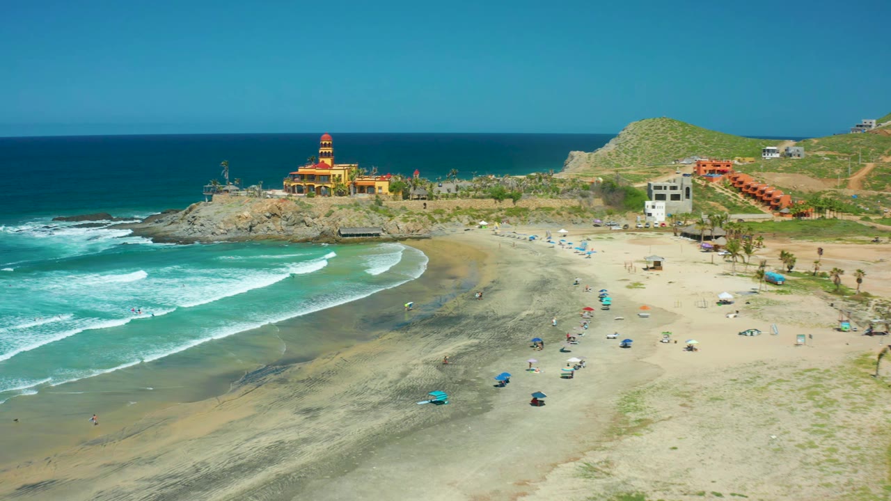 Wide coastal view with surf bands, beach umbrellas, rocky point, and hillside buildings.