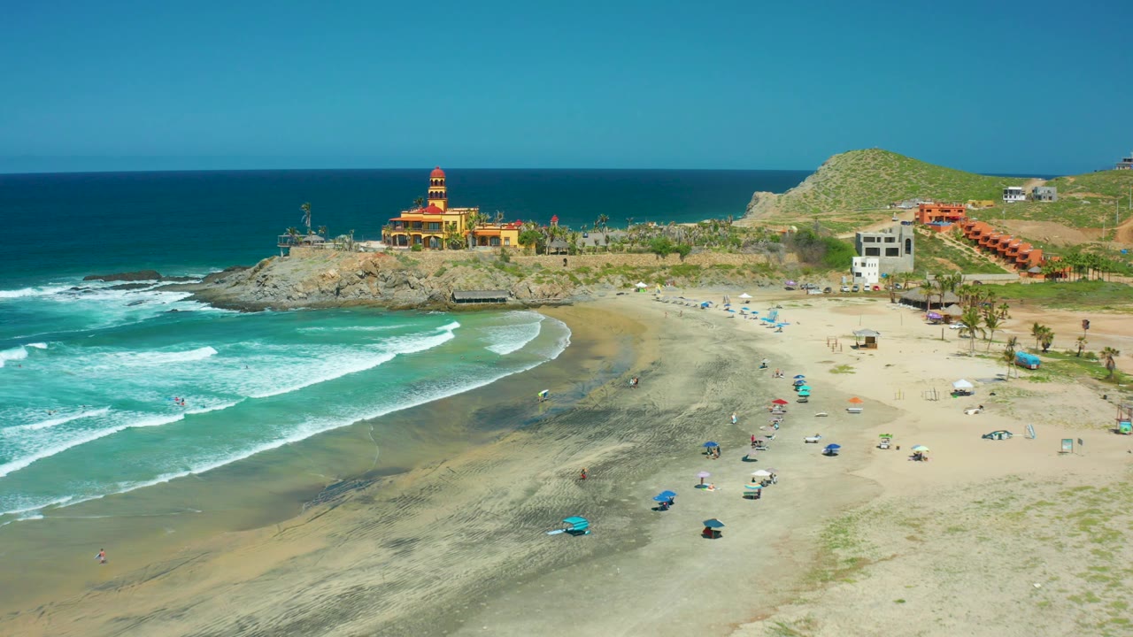 Cerritos Beach seen from an elevated angle, with a larger white wave arc rolling toward the left side of the beach and small umbrellas and people scattered across the open sand.