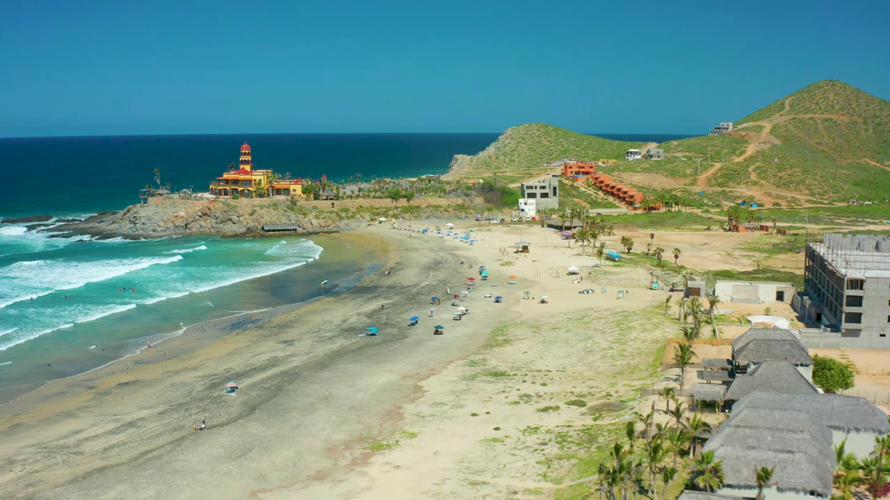 Aerial beach view with waves curving toward umbrellas and buildings near hills.