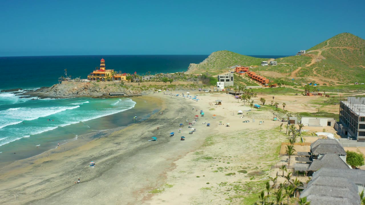 At 0:02, the shoreline view shows turquoise water on the left, pale sand with scattered umbrellas and small figures, palm trees and low buildings on the right, and a green hillside behind them.