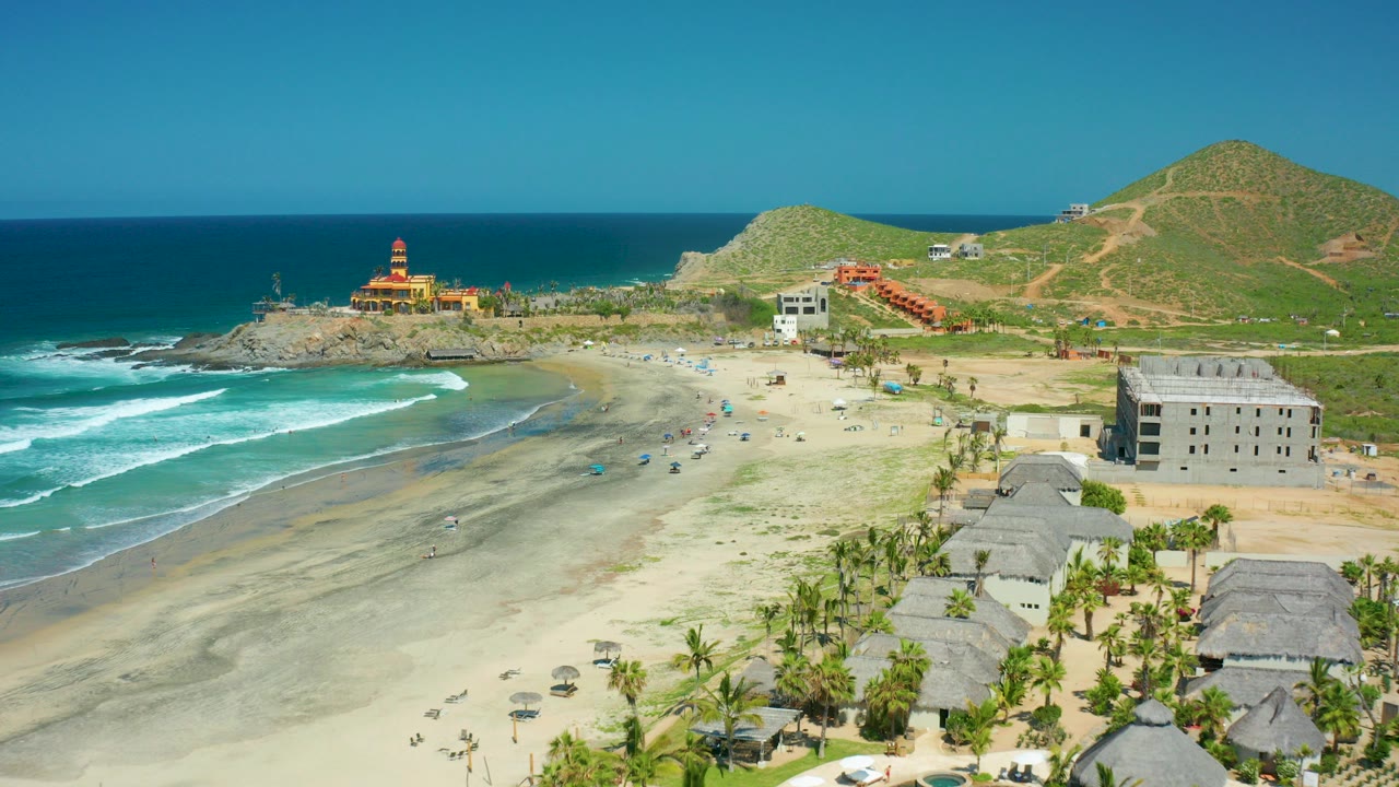 Cerritos Beach aerial frame with surf rolling in from the left, a rocky point and lighthouse-like tower near the water, hillside buildings beyond the sand, thatched huts in the foreground, and no visible people.