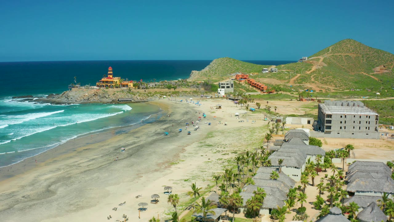 Cerritos Beach frame with deep blue ocean at the horizon, surf breaking at the beach edge, a rocky point with a striped tower, dirt paths on green hills, foreground thatched roofs, and no visible people.