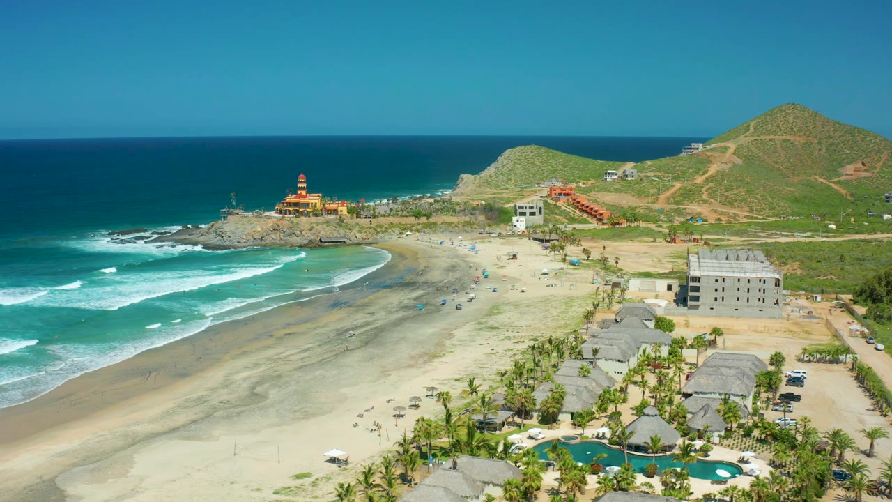 Wide aerial beach view with rolling surf, scattered umbrellas, pool area, hillside, and lighthouse.