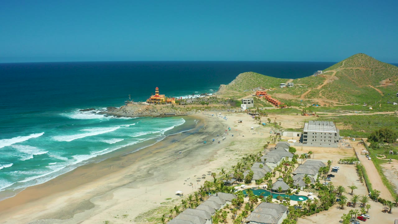 Aerial frame over Cerritos Beach, Baja California Sur, with turquoise waves breaking at left, a wide sand beach, a red-and-yellow lighthouse on the rocky point, shoreline buildings, palms, and green hills behind.