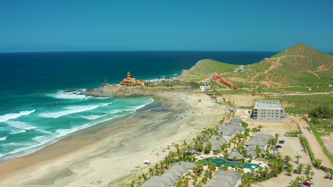 A high drone frame shows Cerritos Beach with layered turquoise water at left, broad pale sand through the center, the lighthouse near the rocky point, and a green hill rising behind scattered shore buildings.
