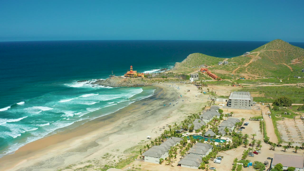 Aerial coastal view with surf, sandy beach, rocky point, buildings, and green hills.