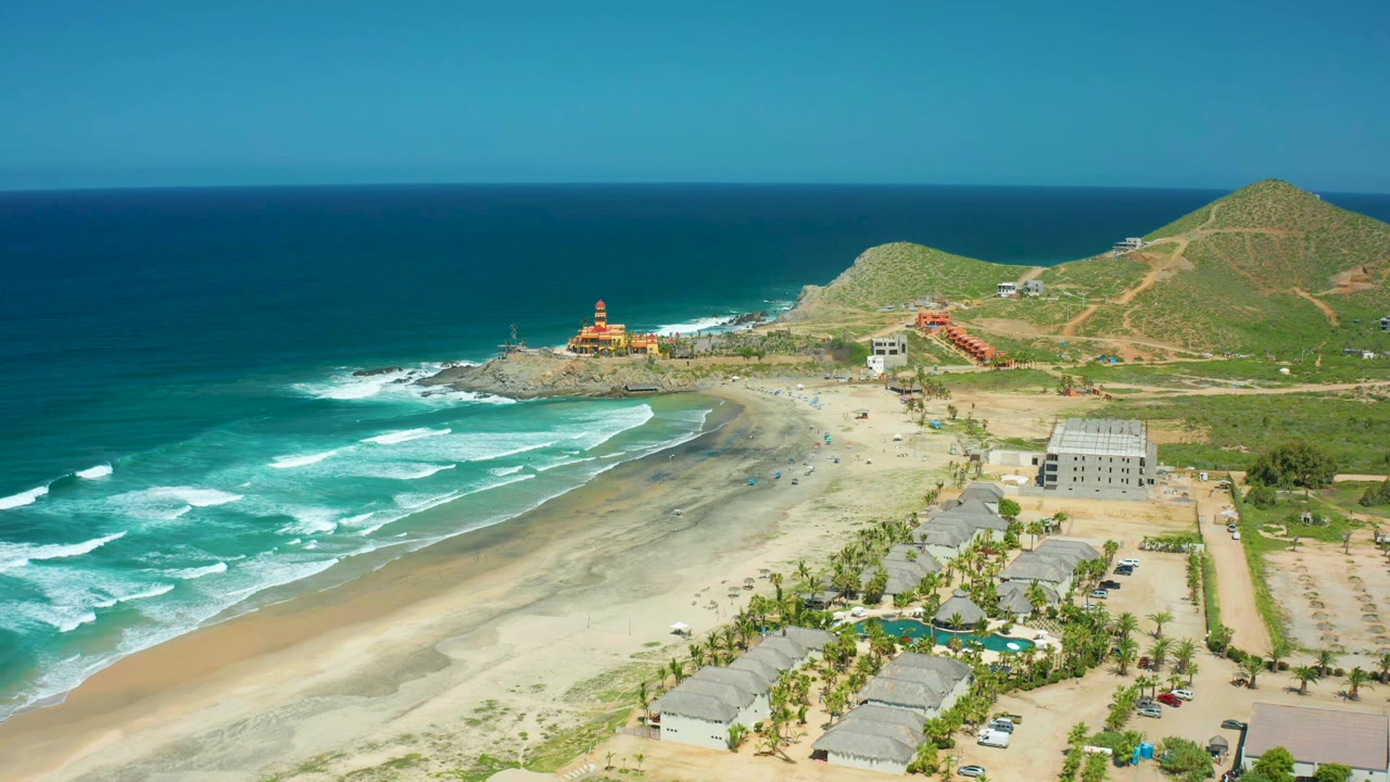 Aerial frame over Cerritos Beach with white wave lines shifting along the left shoreline, a broad sand strip, the rocky point near the center-left, beach buildings, green hills, and no visible people.