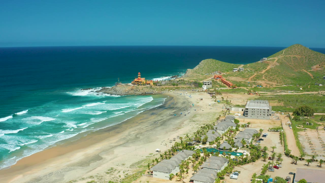 Aerial view of Cerritos Beach showing deeper blue water at the horizon, white surf curling toward the sand, the rocky point, hillside roads, buildings, parked cars, and no visible people.