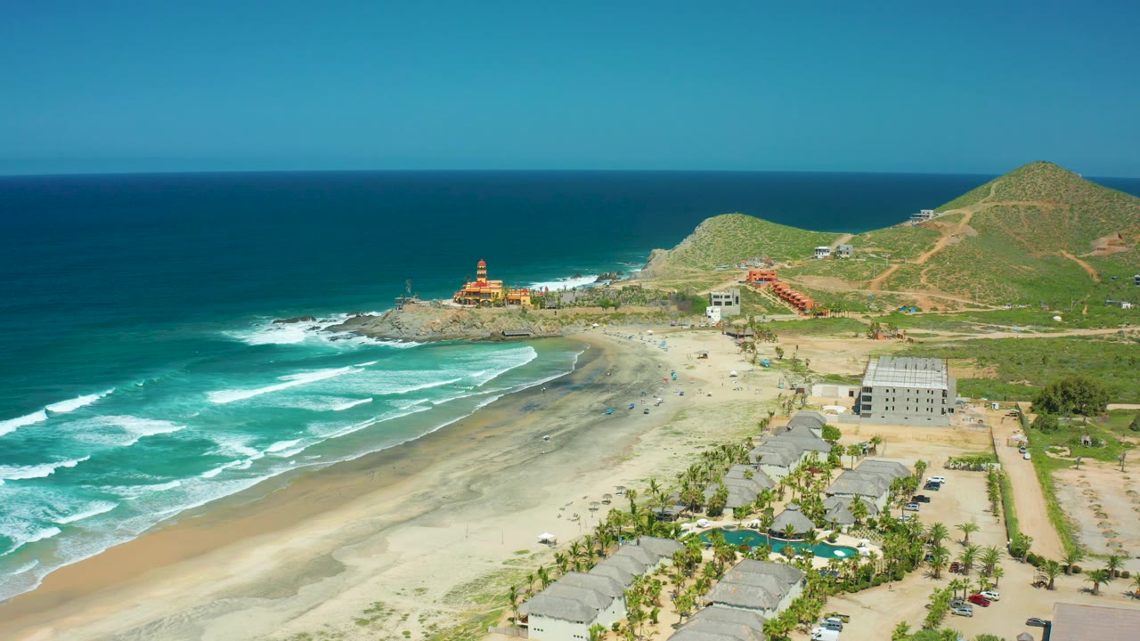 Mid-clip aerial frame of Cerritos Beach with surf bands across the left side, pale sand below, the rocky point and small structure beyond the shore, hillside roads, buildings, cars, and no visible people.