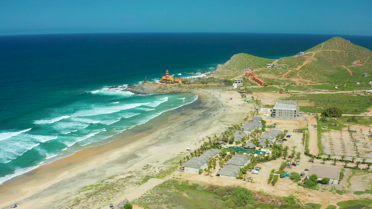At 0:01, the aerial view over Cerritos Beach shows surf bands closer to shore, light sand, palm-lined buildings, hillside roads, the red crane or structure, and no visible people.