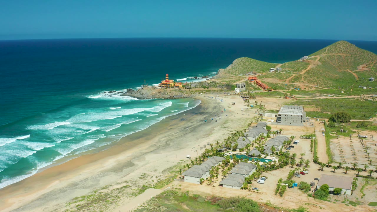 At 0:03, the frame shows Cerritos Beach with rolling waves on the left, sand and low structures in the center, parking rows to the right, green hills behind, and no visible people.