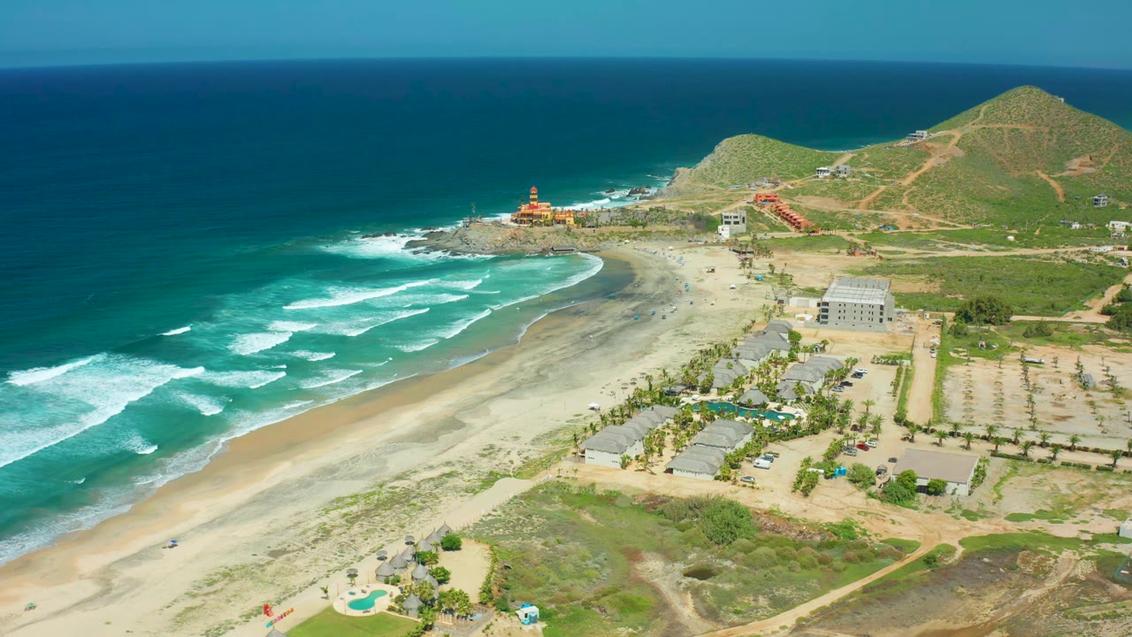 Cerritos Beach aerial frame one second in, showing white wave bands closer to shore, beach sand, scattered buildings, dirt tracks, green hills, and no visible people.
