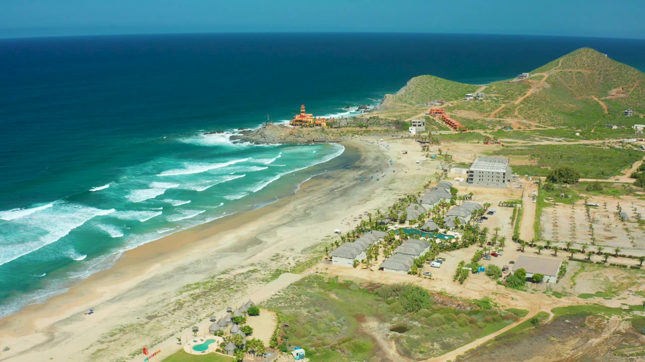 Cerritos Beach aerial frame with a broad sweep of turquoise water, curving surf, sandy shore, low structures, hillside paths, and no visible people.