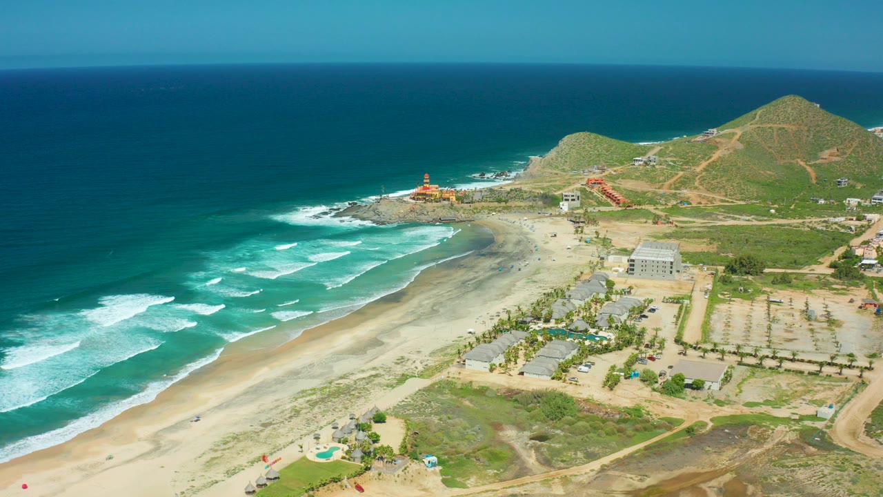 Aerial frame of Cerritos Beach in Baja California Sur with teal waves reaching the sandy shore, low green hills beyond, scattered buildings, and no visible people.