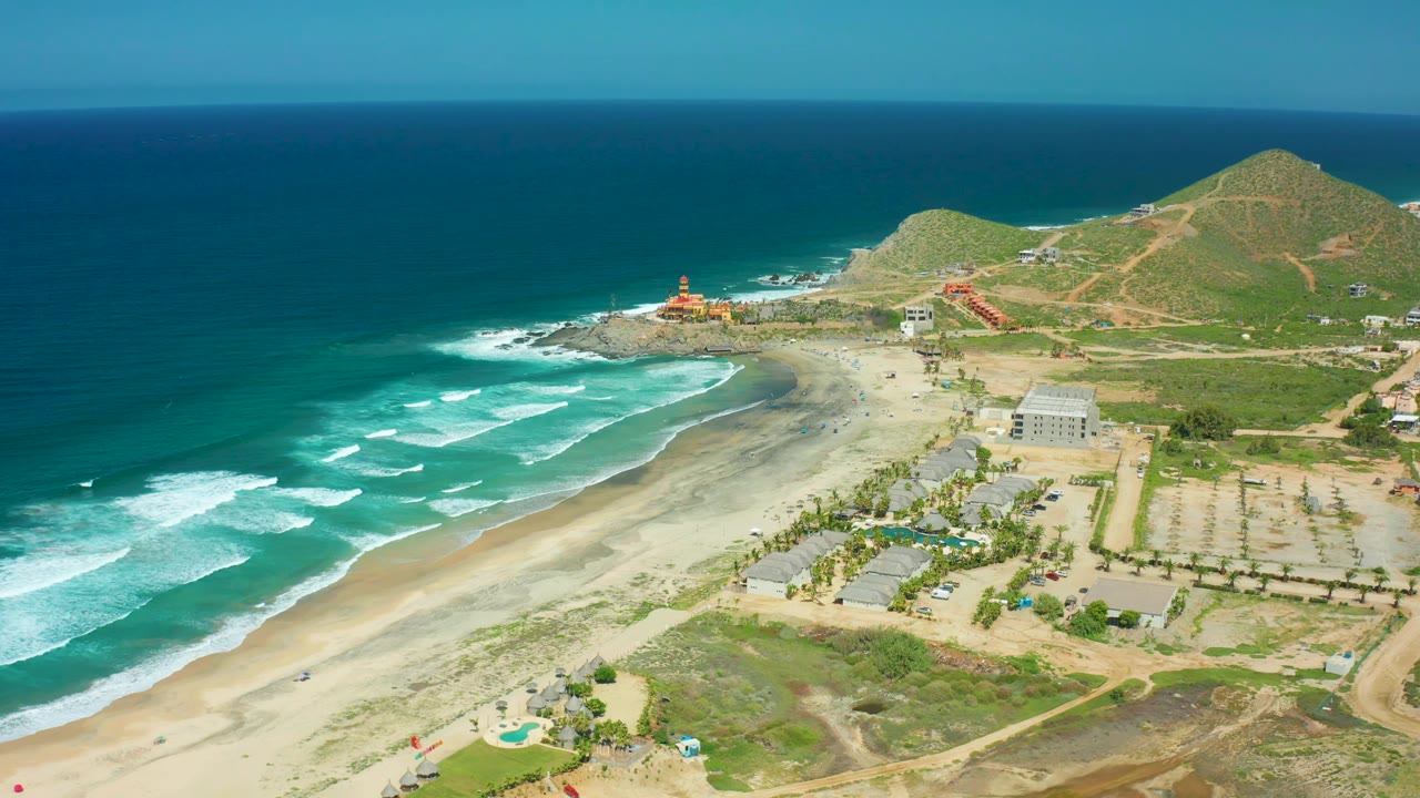 Aerial frame over Cerritos Beach showing turquoise surf, broad sand, a few beachside buildings, hillside paths, deep blue ocean, and no visible people.