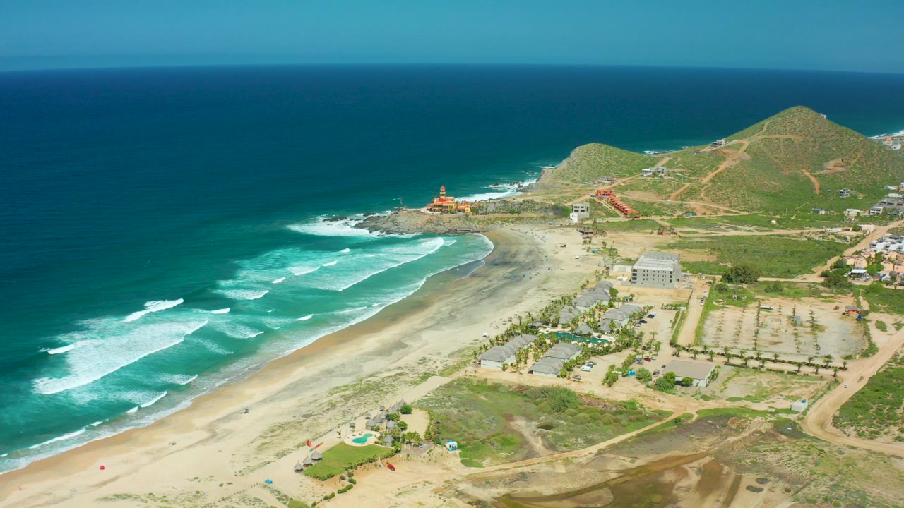 Opening aerial frame over Cerritos Beach in Baja California Sur, Mexico, showing teal waves, white foam, sandy shoreline, a lighthouse on rocks, low buildings, parking areas, green hills, and no visible people.