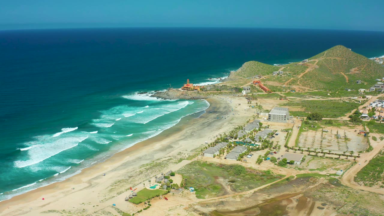 Aerial frame at Cerritos Beach with broad bands of white surf moving toward sand, a lighthouse near the rocky point, clustered beach structures, parking rows, dirt tracks, green hills, and no visible people.