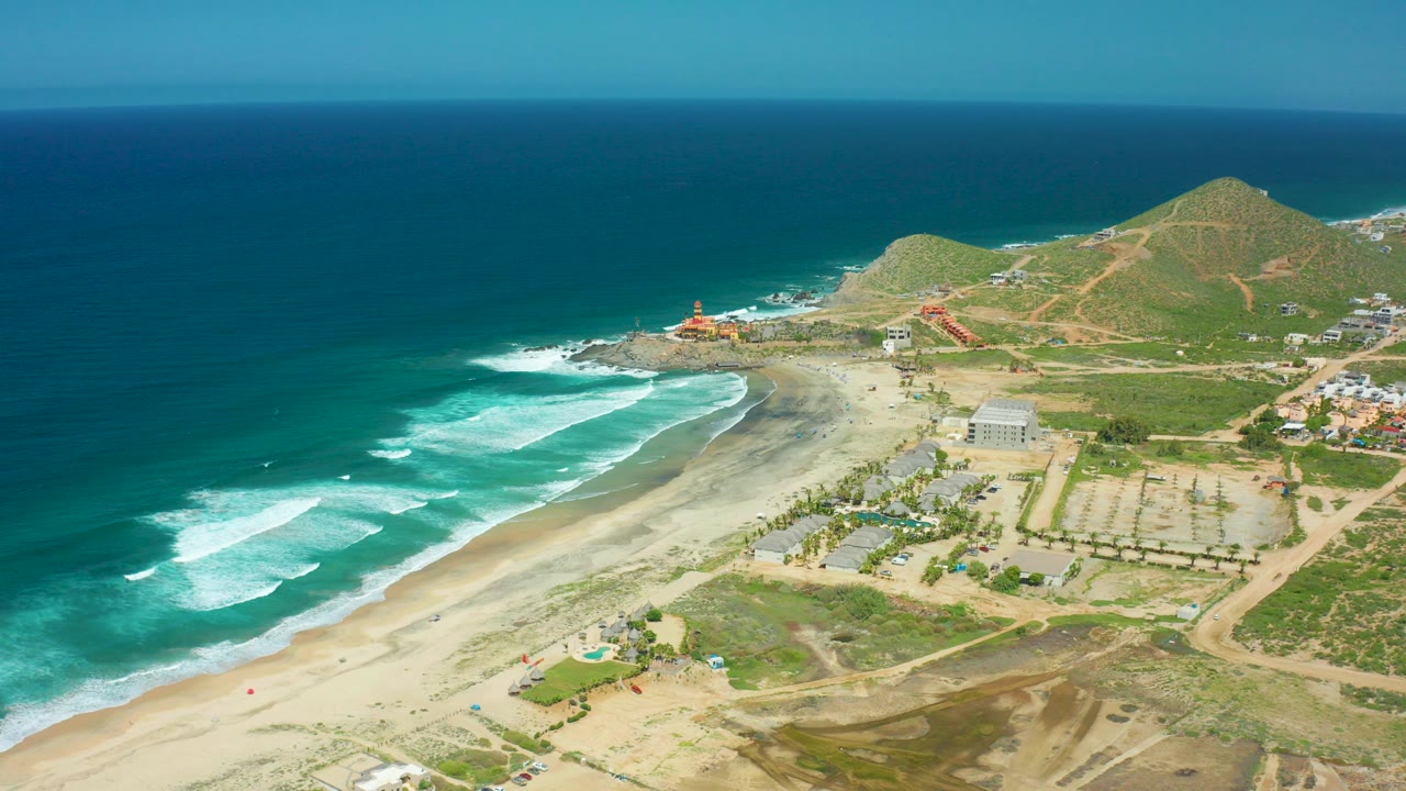 Aerial view over Cerritos Beach in Baja California Sur, Mexico, with turquoise surf on the left, sandy shoreline below, scattered coastal buildings inland, green hills beyond, and no visible people.
