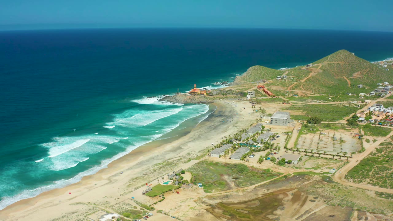 Wide aerial frame of Cerritos Beach, Baja California Sur, Mexico, showing fresh white wave bands near shore, a pale sand beach, low buildings and roads inland, green hills in the distance, and no visible people.