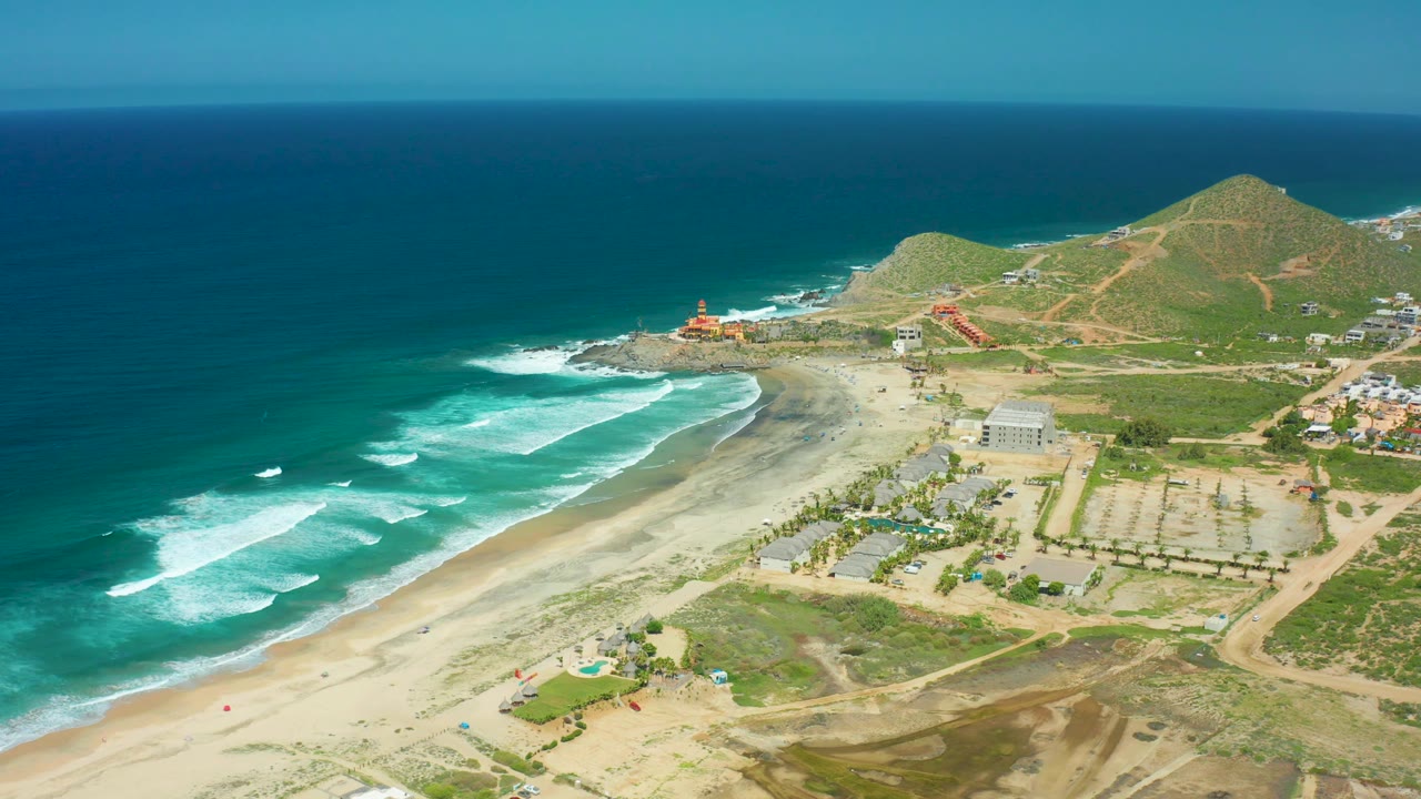 Aerial coastal view with waves, sandy beach, palms, buildings, roads, and hills.
