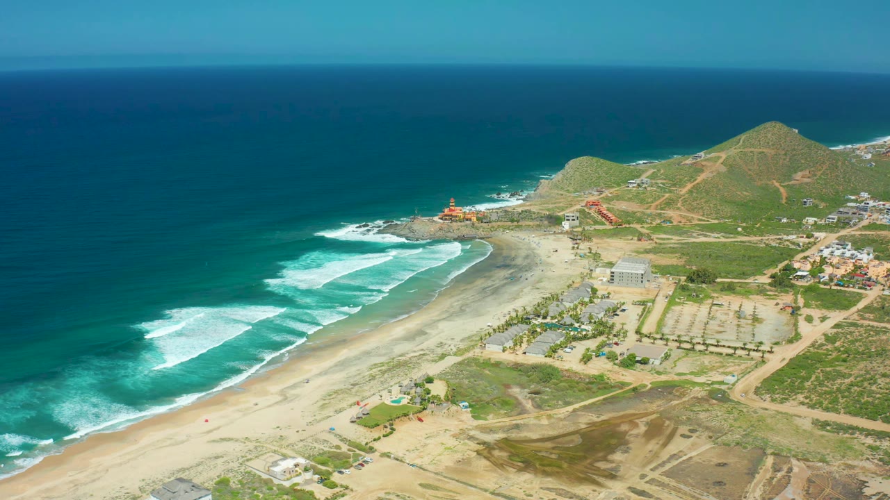 Aerial view at Cerritos Beach with parallel white wave lines approaching the sand, darker open water offshore, inland roads, low buildings, and no recognizable people.