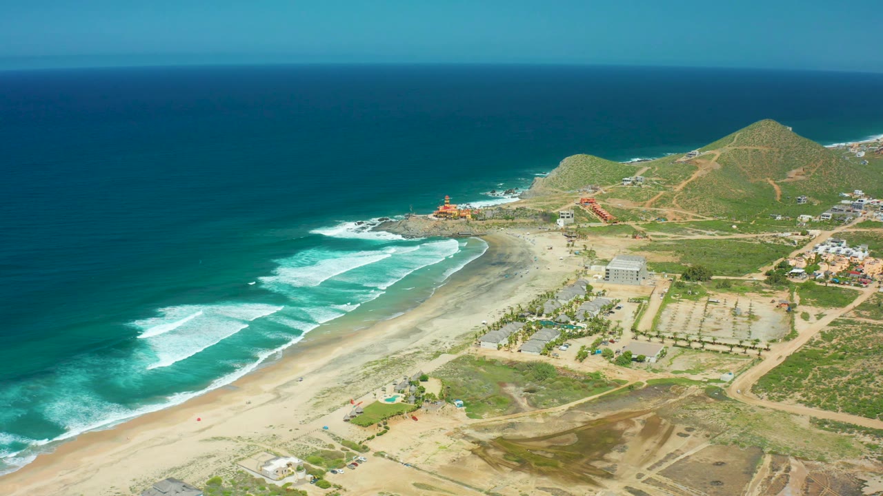 High oblique aerial frame of Cerritos Beach showing surf curling toward the beach, pale sand, a cluster of inland buildings, dirt tracks, a rounded hill, and no recognizable people.