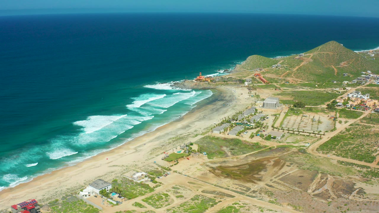 Opening aerial frame over Cerritos Beach, Baja California Sur, with blue ocean, white surf, sandy shore, low hills, dirt roads, scattered buildings, and no visible people.