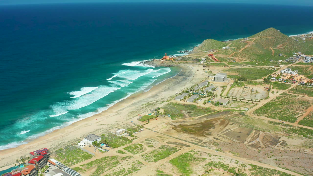 Cerritos Beach coastline from above, showing waves curling toward the sand, a pale beach, dirt access roads, low structures, green hillside terrain, and no visible people.