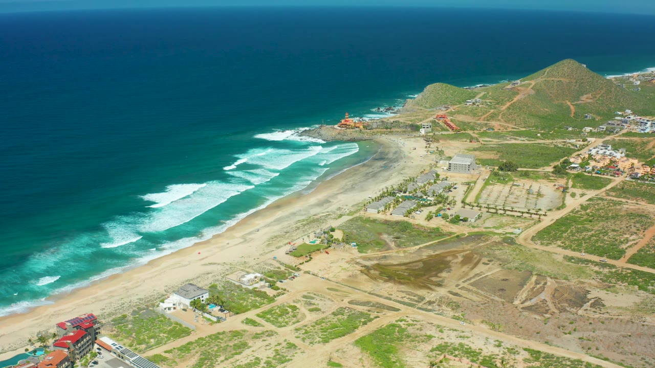 High view of Cerritos Beach with deep blue water, white wave sets, sandy shoreline, inland dirt tracks, small coastal buildings, rounded hills, and no visible people.