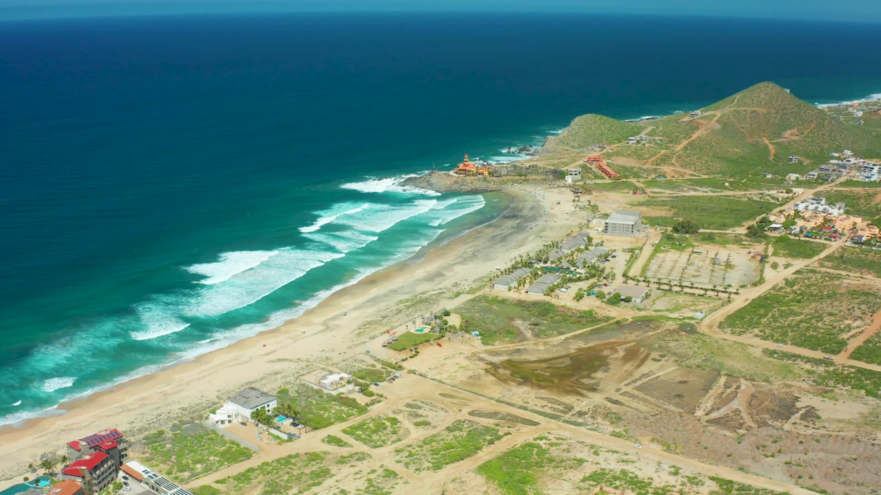 Aerial frame of Cerritos Beach where white foam pushes across the shoreline, with sandy flats, dirt roads, green scrub, low buildings, hills, and no visible people.