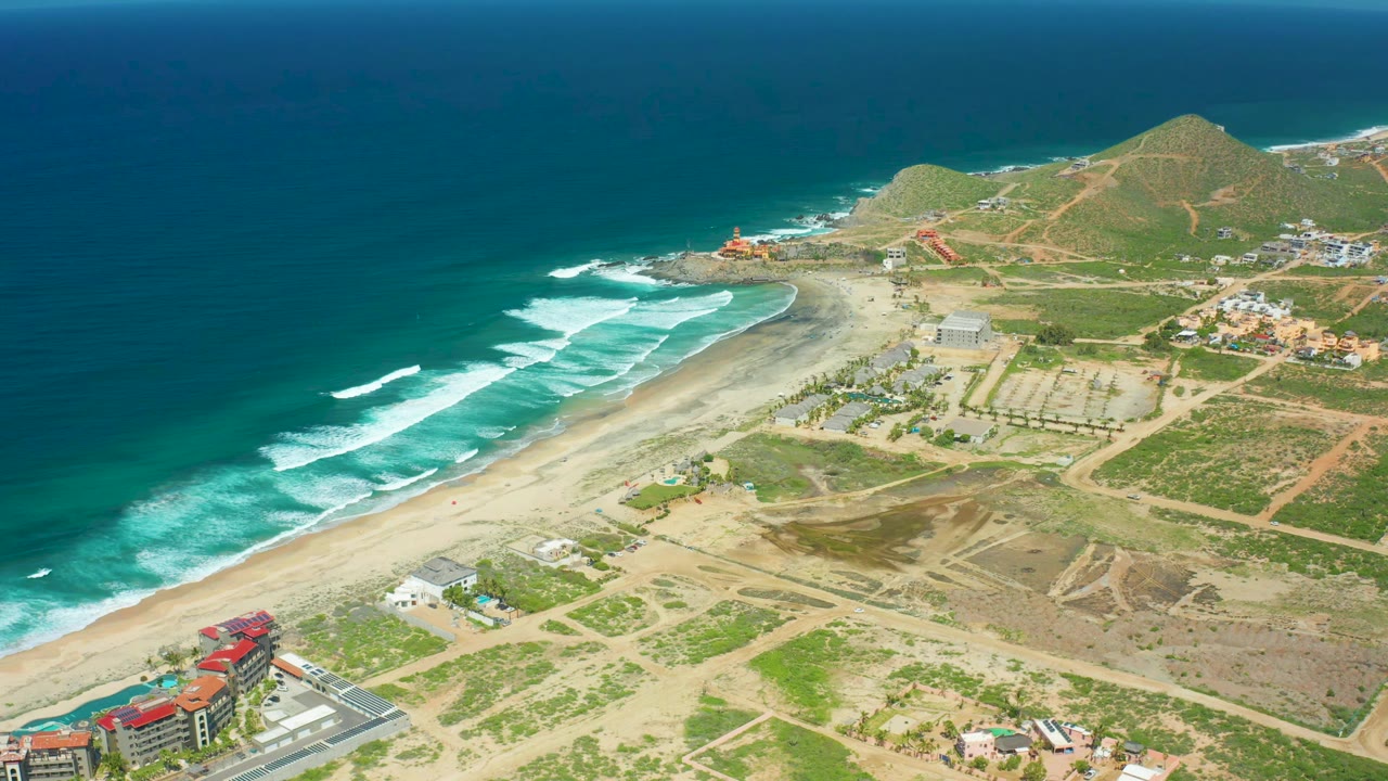 Aerial frame of Cerritos Beach showing turquoise water, white wave breaks, pale sand, low green hills, dirt tracks, coastal buildings, and no visible people.