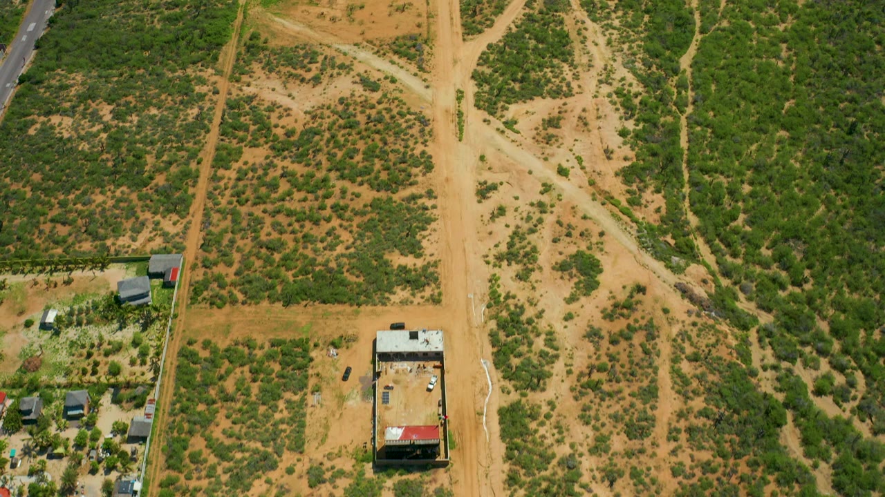High aerial view near Cerritos Beach, Baja California Sur, with the dirt-road crossing centered, dry green patches around it, a small building low in frame, and no visible people.