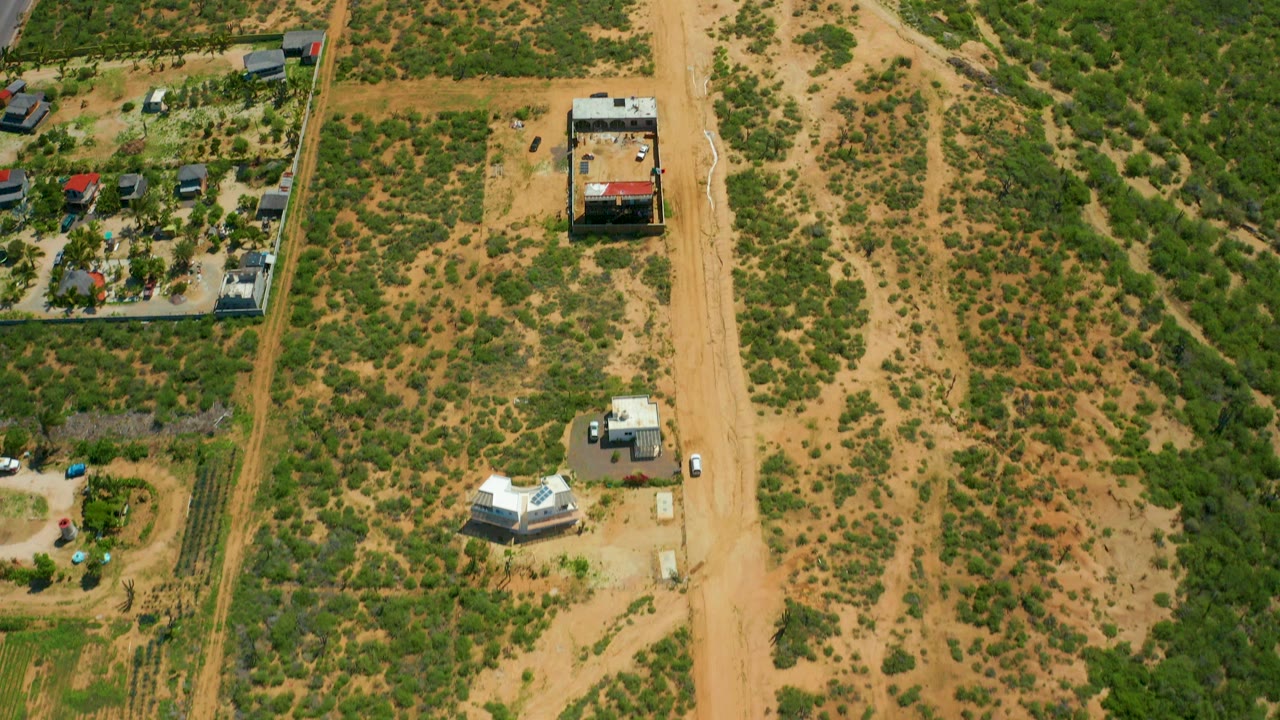 Overhead view near Cerritos Beach, Baja California Sur, showing a sandy dirt road, green desert shrubs, a white structure, a red-roofed building, parked cars, and no visible people.