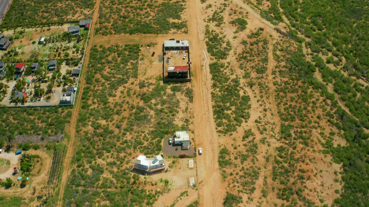 Drone-style overhead view near Cerritos Beach, Baja California Sur, showing a straight sandy road, patchy green vegetation, small buildings, parked vehicles, and no visible people.