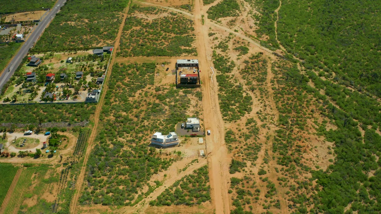 Aerial frame over Cerritos Beach, Baja California Sur showing a sandy dirt road, small rural structures, planted lots, green scrub, tan open ground, and no visible people.