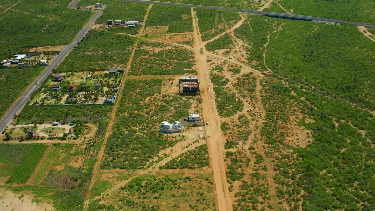 Aerial frame over Cerritos Beach, Baja California Sur, Mexico, with a paved road along the left edge, tan dirt tracks crossing green scrubland, scattered rural buildings, and no visible people.