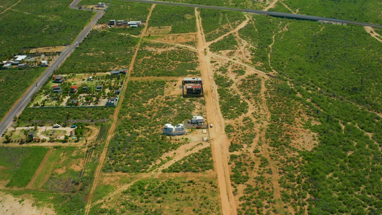 Aerial view in Cerritos Beach, Baja California Sur, Mexico, showing the paved road slightly farther left, a central dirt road, patchy green fields, several rural structures, and no visible people.