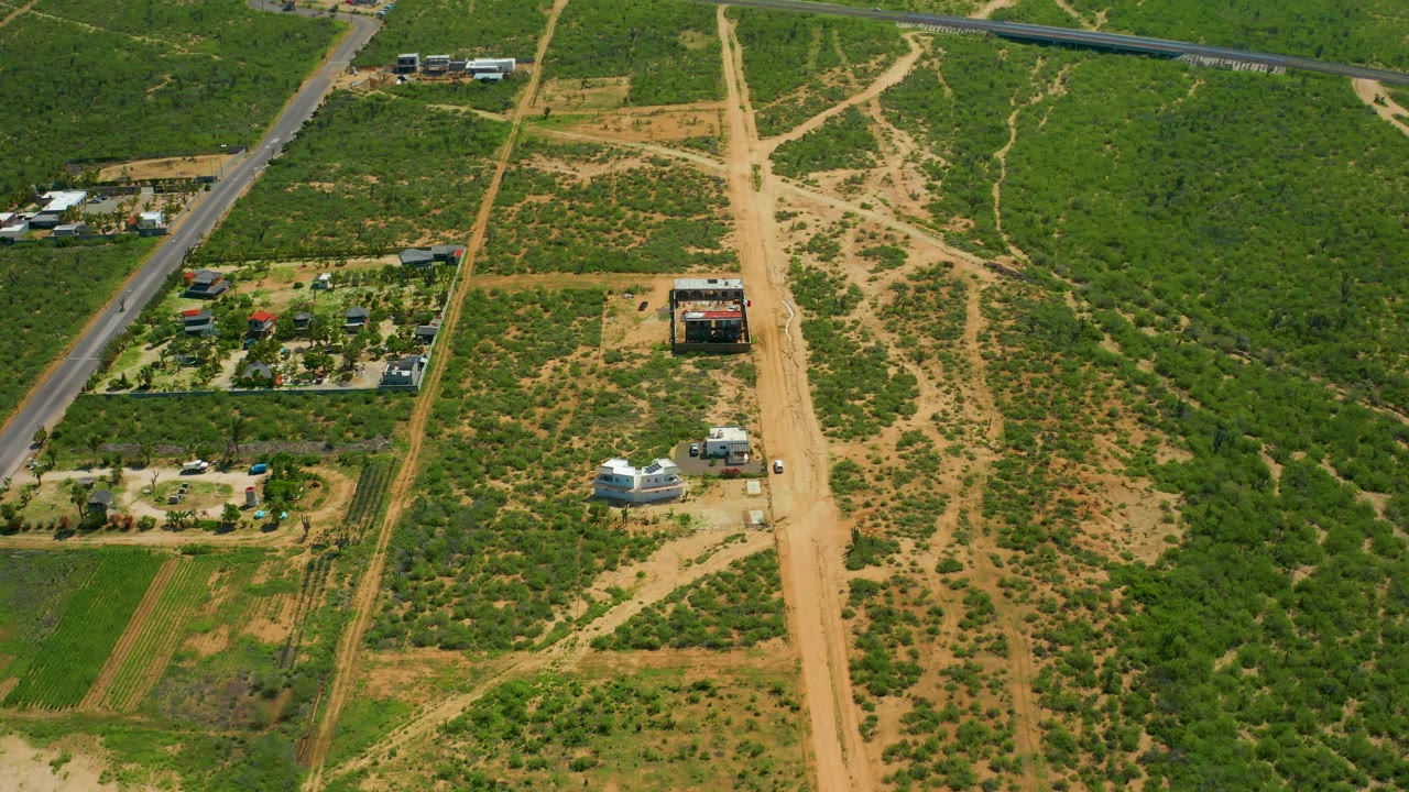 Drone-view frame over Cerritos Beach, Baja California Sur, Mexico, with the paved road at left, a broad tan dirt track near center, low green vegetation, rural structures, and no visible people.