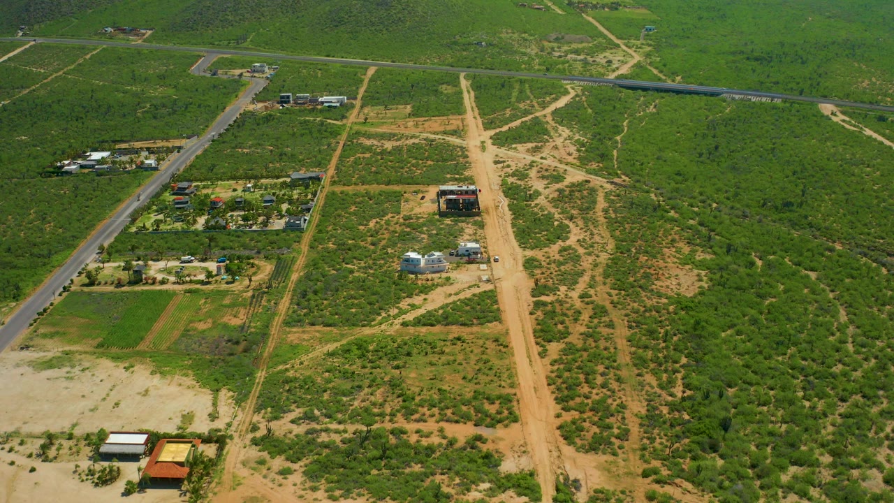 Opening aerial frame over Cerritos Beach, Baja California Sur shows a paved road beside green fields, sandy dirt roads, farm buildings, and no visible people.