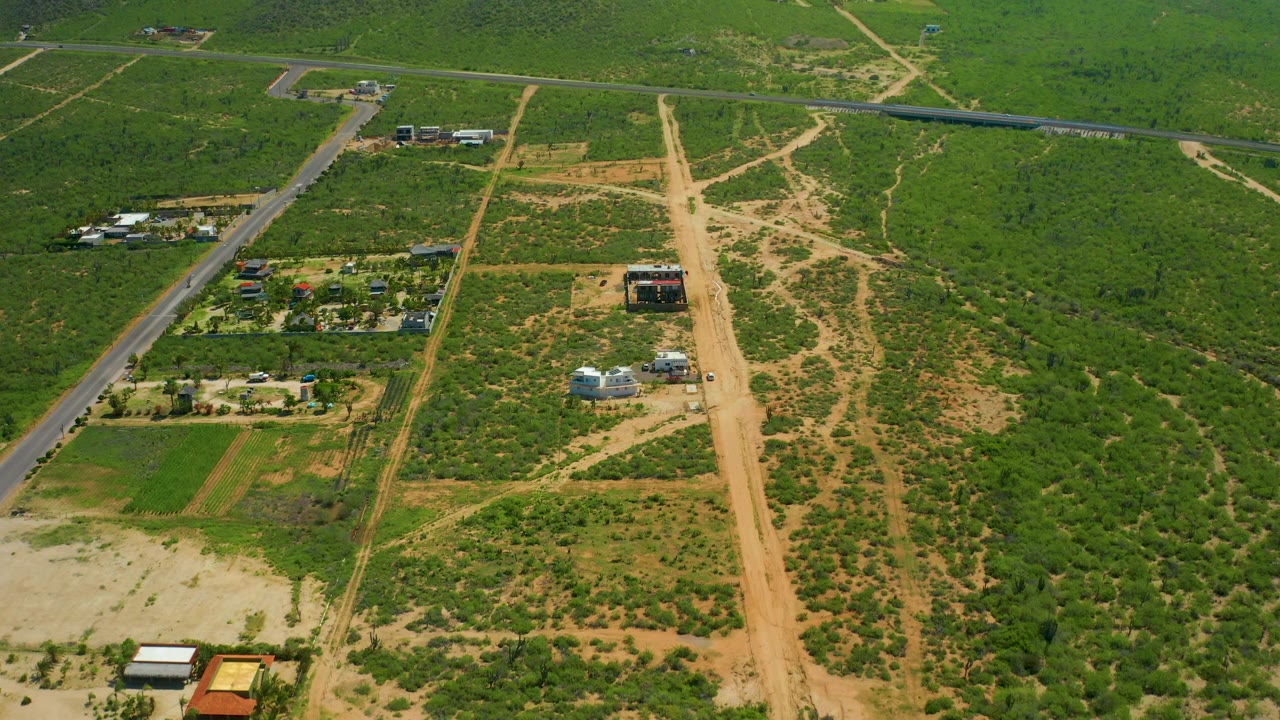 At 0:02, a high aerial frame over Cerritos Beach shows the paved road, nearby farm compounds, tan dirt routes, green vegetation, and no visible people.