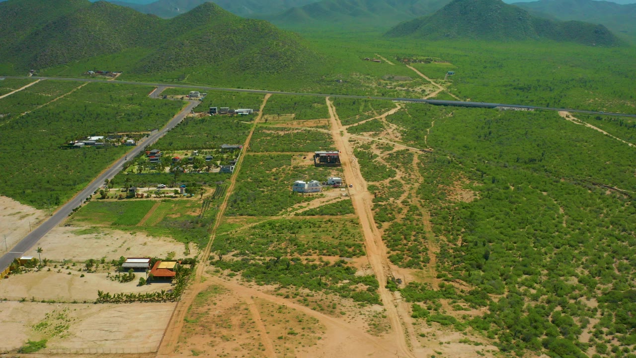 Aerial view over Cerritos Beach, Baja California Sur, showing a paved road at left, a central dirt track, scattered small buildings, green vegetation, distant hills, and no visible people.