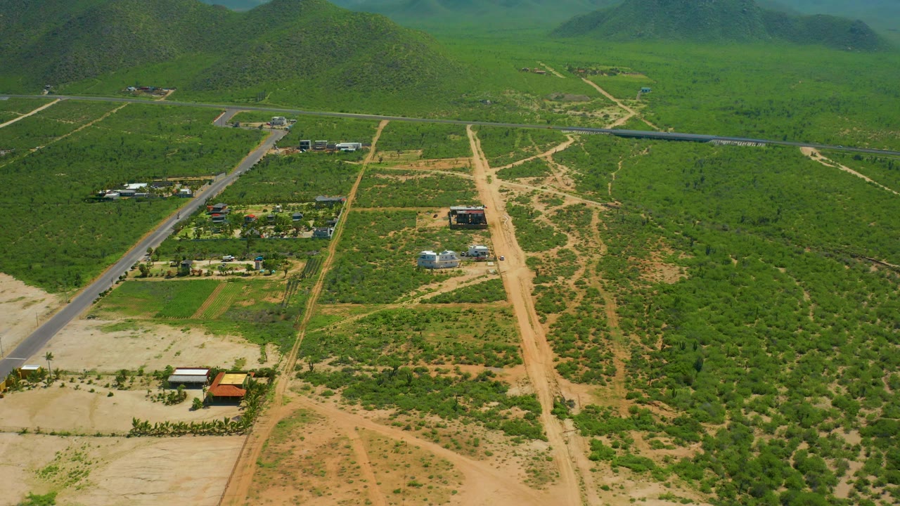 Drone frame over Cerritos Beach, Baja California Sur, showing branching dirt tracks, a paved road, clusters of small buildings, green vegetation, rounded background hills, and no visible people.