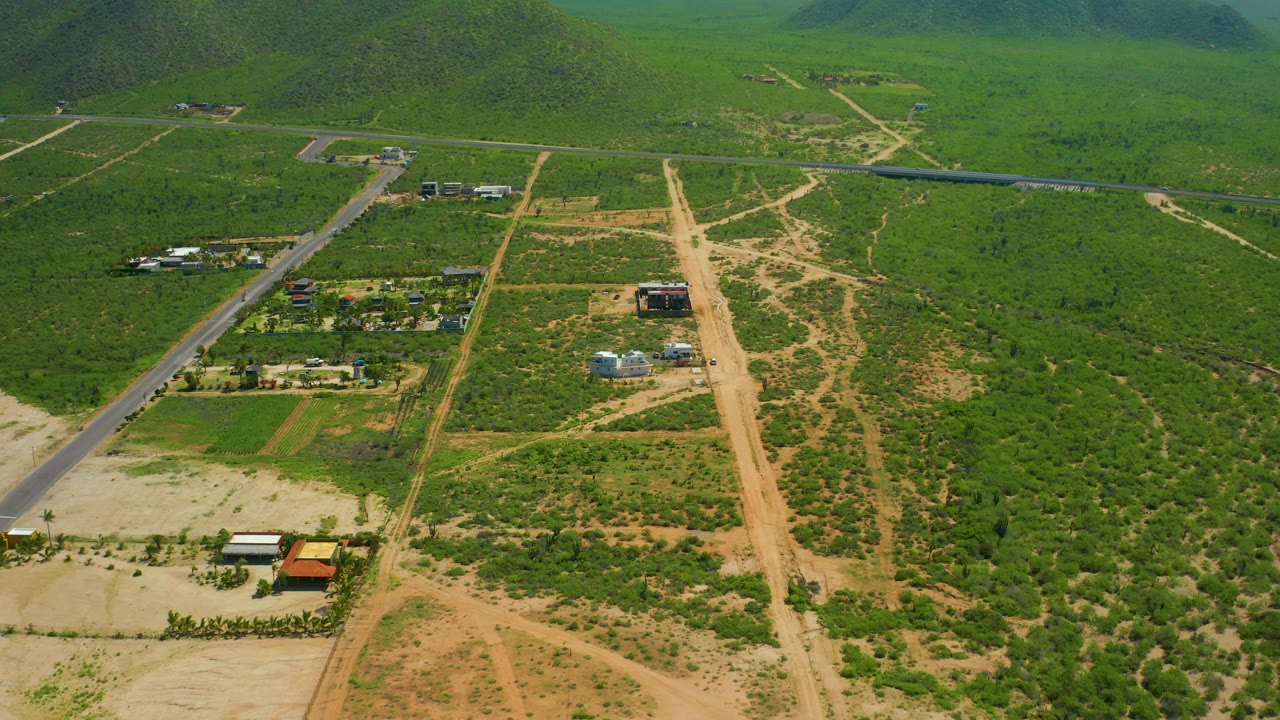 Rural aerial view with a paved road, tan dirt road, buildings, fields, and hills.