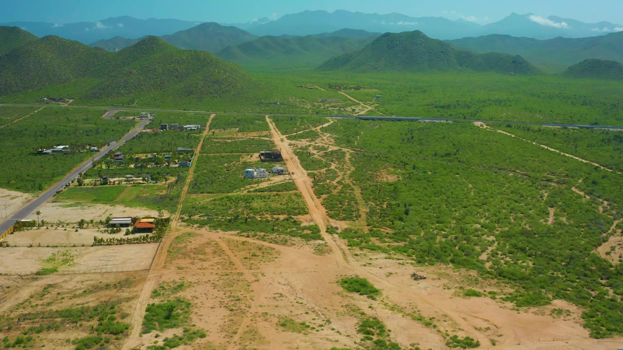 Drone view across rural roads, scattered buildings, green vegetation, hills, and mountains.
