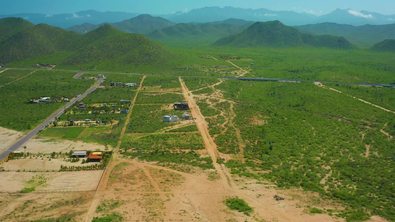 Aerial frame of a rural road, open lots, buildings, green hills, and distant mountains.
